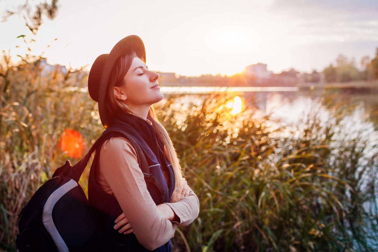 Woman with backpack and hat next to lake taking a deep breath